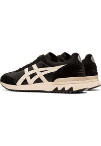 Black athletic sneakers with beige accents, suede and nylon materials, rounded toe, textured sole, and distinctive side logo stripes.