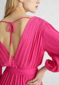 Abito plissettato fucsia con profondo scollo a V sul retro, sottili spalline regolabili e un nastro annodato con un elegante tassello dorato sul retro.