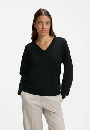 Femme aux cheveux bruns et raides portant un pull noir à col en V et un pantalon beige, debout les mains dans les poches sur un fond uni.