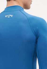 Zils garas piedurknes rash guard, izgatavots no gludas, elastīgas auduma. Iespējas plakanās šuves un neliels balts logo augšējā muguras daļā.