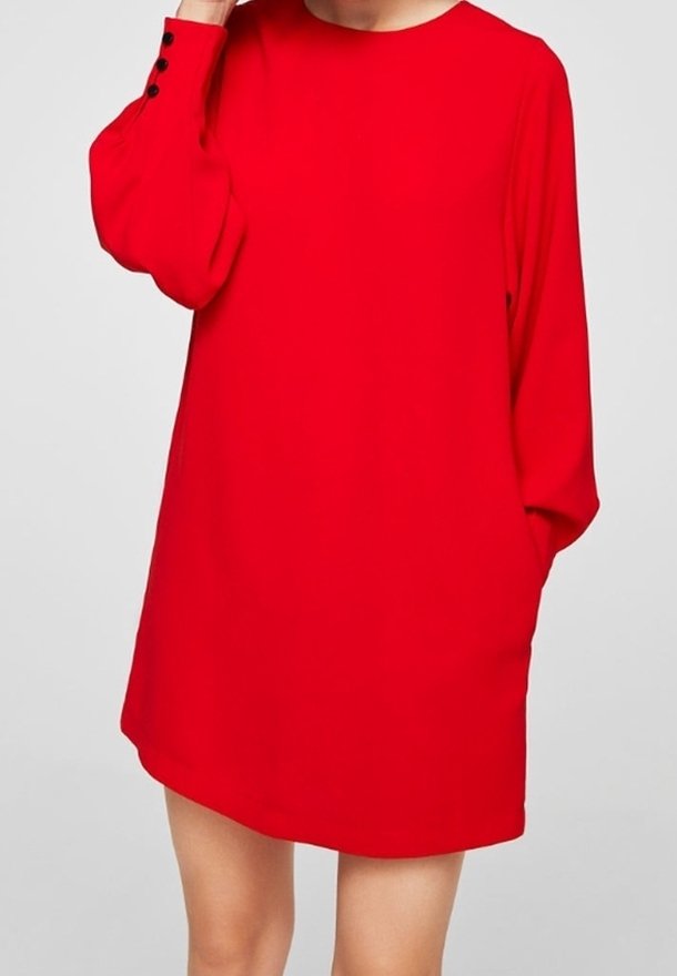 Femme portant une robe rouge ample à manches longues avec des détails de boutons sur un poignet, debout devant un fond clair uni.