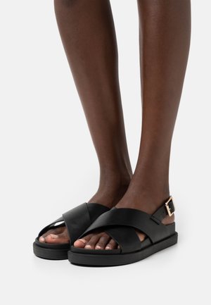 Black leather sandals featuring cross-strap design, adjustable buckle, flat sole, and exposed toes. Smooth texture, minimalistic style.