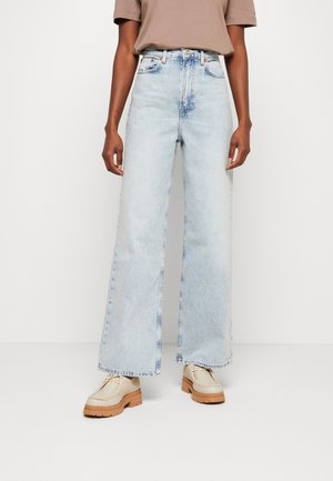 Person trägt hellblaue, hoch taillierte Jeans mit weitem Bein, beigefarbene Schuhe mit dicker Sohle und ein in die Hose gestecktes, kurzärmeliges Taupe-Oberteil und steht vor weißem Hintergrund.