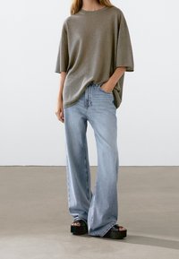 Ylimittaisen suuri oliivinvihreä puuvillainen t-paita, jossa on lyhyet hihat, yhdistetty vaaleansinisiin leveälahkeisiin farkkuihin. Mustat korolliset sandaalit viimeistelevät asun.