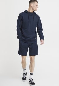 Jūras spēku kapuce ar priekšējo kabatu un regulējamu auklu, apvienota ar saskaņotām šortiem. Valkāta ar melniem sporta apaviem un baltām sporta zeķēm.