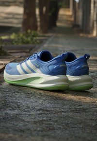 Chaussures de course bleues avec des bandes blanches et des semelles vert clair posées sur un chemin de pierre à l'extérieur avec des arbres flous en arrière-plan.