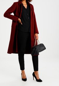 Cappotto bordeaux con colletto, blusa nera e pantaloni neri aderenti. Accessorio: piccola borsa nera con frange. Tacchi neri completano il look.