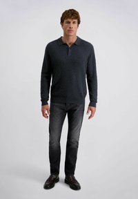 Man staat in een donker gestructureerd langpoloshirt, vervaagde zwarte jeans en donkerbruine leren instappers op een effen achtergrond.