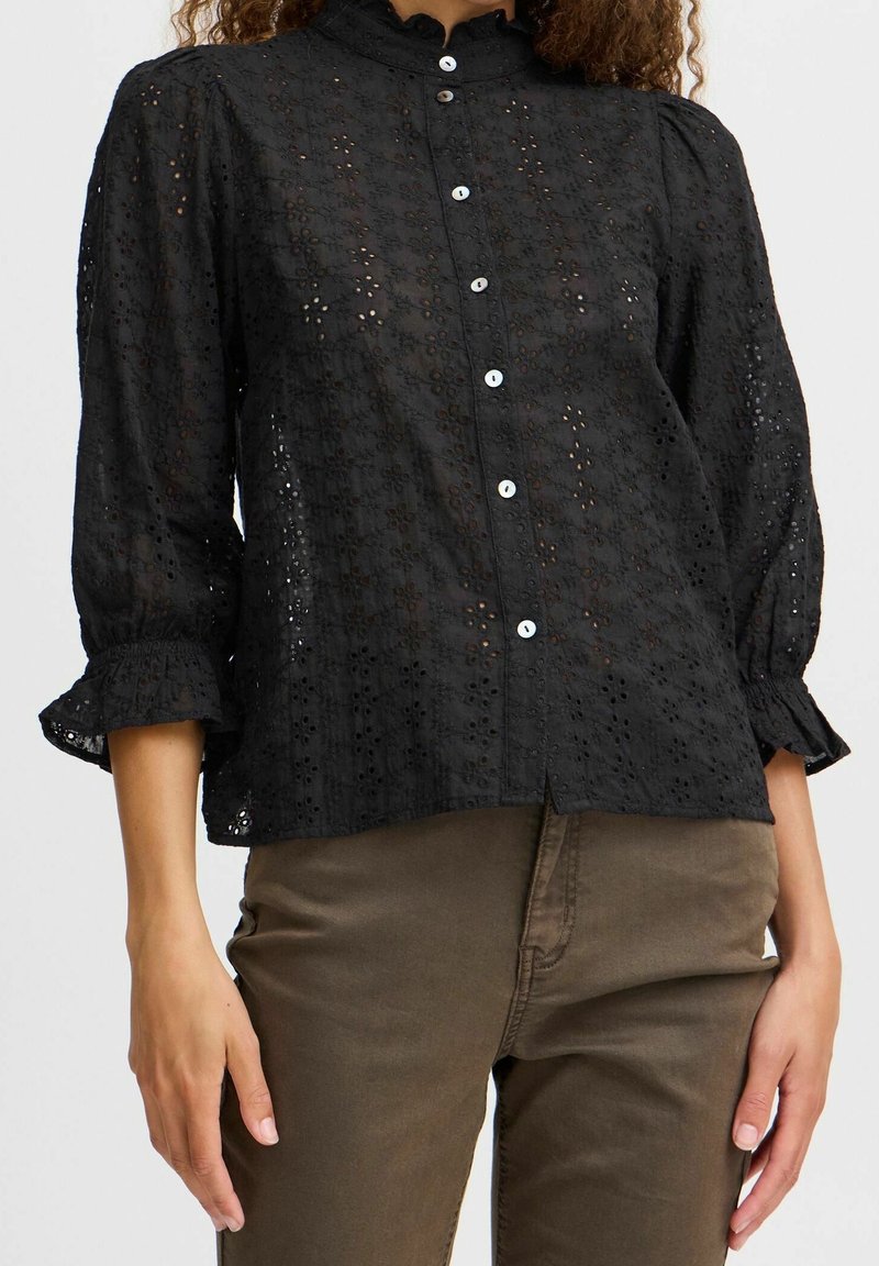 Blouse en dentelle noire à œillets avec des boutons blancs, manches trois-quarts à volants, associée à un pantalon vert olive.