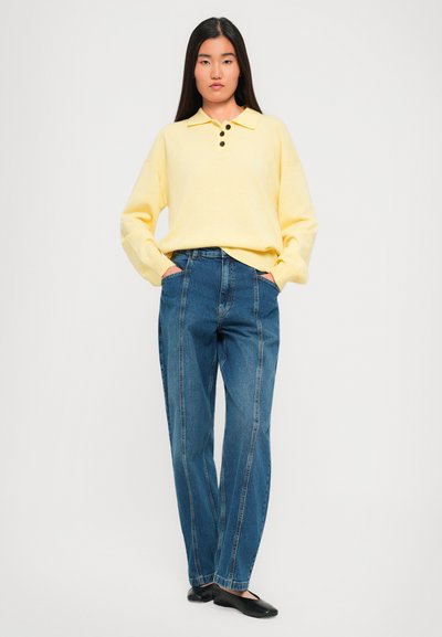 Woman standing with hands in pockets wearing a pale yellow long-sleeve collared sweater, high-waisted blue jeans, and black flats.