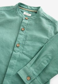 Chemise verte à manches longues avec col mandarin, boutons en bois, poche poitrine et manches retroussées, posée à plat sur une surface blanche.