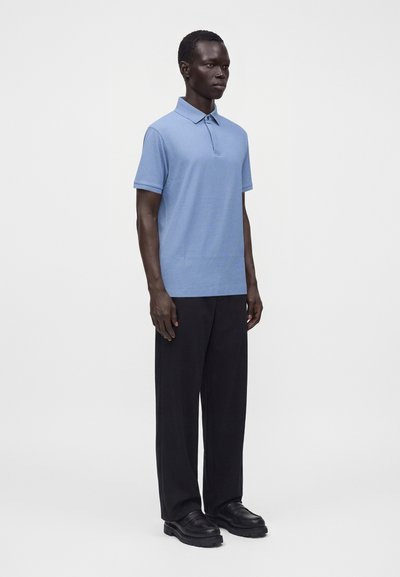 Homme portant un polo bleu clair à manches courtes, un pantalon noir à jambes larges et des chaussures noires, debout devant un fond blanc uni.
