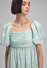 Vaaleansininen mekko, jossa on neliökaula-aukko, puffihihat ja kerrostettu pinta. Kankaan kukkakuviot ja virtaava siluetti.