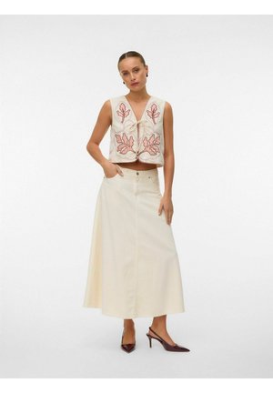 Femme portant un gilet crème sans manches avec une broderie de feuilles roses, une longue jupe crème et des chaussures à talons slingback marron foncé, debout les mains dans les poches.