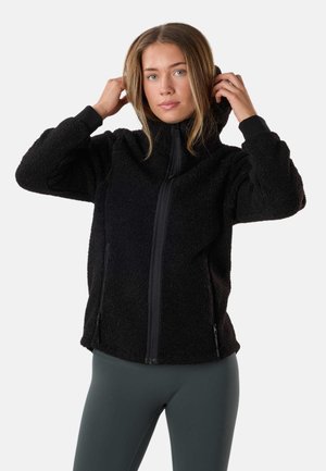 Kvinde, der justerer hætten på en sort fleecejakke med lynlås foran, iført mørke leggings, vendt mod fronten på en ensfarvet lys baggrund.