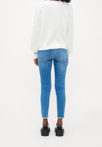 Biały sweter z teksturowanymi rękawami, niebieskie wąskie dżinsy i czarne buty na platformie. Cechy to zaokrąglony dekolt i postrzępione wykończenia.