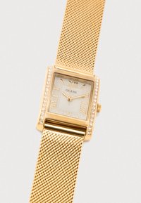 Gouden horloge met metalen meshband en een vierkante wijzerplaat, met heldere kristalaccenten en Romeinse cijfers. De wijzerplaat is crème met geometrische patronen.