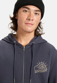 Navy zip-up hoodie made from soft fabric, featuring a front pocket, drawstrings, and a beige sun graphic logo on the left chest.