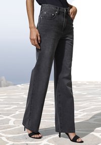 Jeans grigi scuri a vita alta e gamba larga, dotati di tasche frontali, dettagli cuciti e accessori in argento; abbinati a sandali neri con tacco.