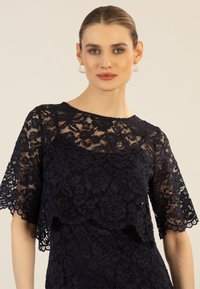 Marinebluse aus Spitze mit kurzen, weiten Ärmeln, verziert mit floralen Mustern und einem gewellten Saum. Die transparente Texturüberlagerung verleiht Tiefe und Detail.