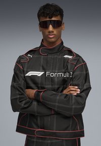 Black racing jacket with white grid lines and red accents. Features a high collar and logo on the chest. Model wears sunglasses.