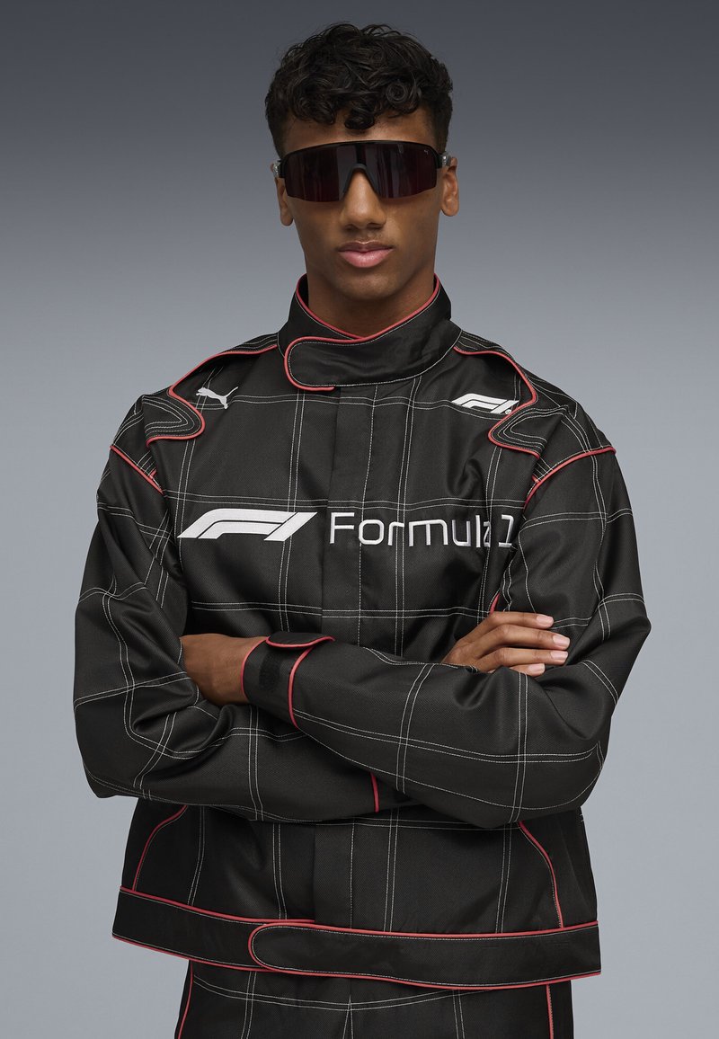 Black racing jacket with white grid lines and red accents. Features a high collar and logo on the chest. Model wears sunglasses.