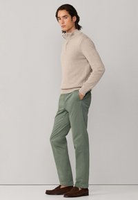 Beige trui met rits aan de hals, groene broek en bruine suède loafers. De trui heeft een gestructureerde afwerking; de broek heeft zakken en een getailleerde pasvorm.