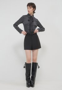 Schwarze Bluse mit Polka-Dots und Schleife, lange Ärmel, kombiniert mit hochgeschnittenen schwarzen Shorts. Glänzende kniehohe schwarze Stiefel mit Quasten.