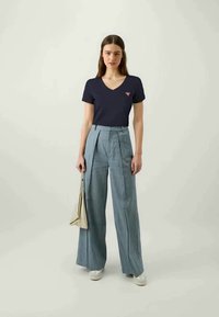 Navy v-neck t-shirt with small red logo, paired with high-waisted, wide-leg light denim trousers and white shoes. Holding a beige bag.