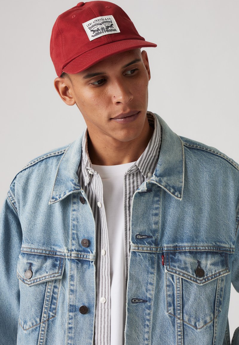 Rød baseballcap med designpatch, lys blå denimjakke med lommer og en grå stripete skjorte under, med hvit rund hals.