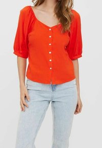 Blouse rouge à boutons avec des manches bouffantes, en tissu léger, design en V, et boutons blancs sur le devant. Associée à un jean bleu clair.