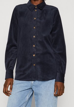 Personne portant une chemise en velours côtelé bleu marine à boutons avec des manches longues et un jean en denim bleu clair, debout devant un fond uni.