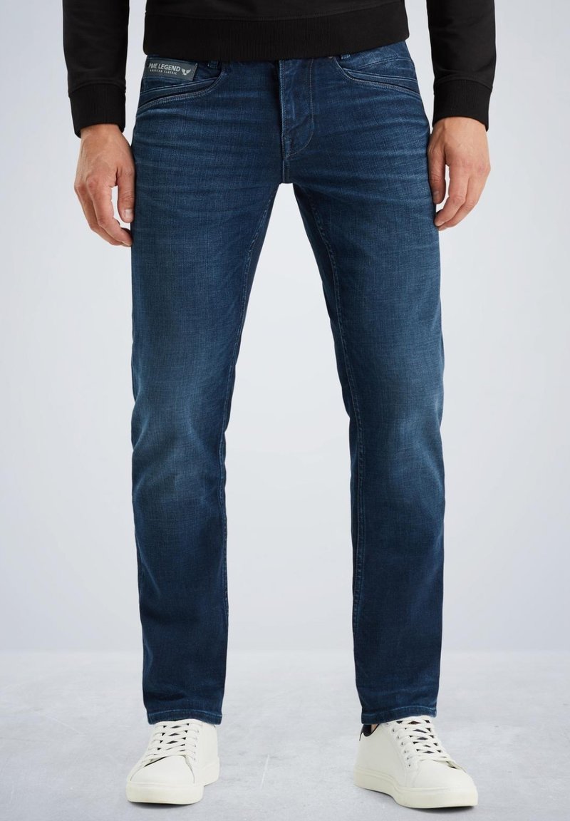 Jeans oscuros de corte slim, con un leve desgaste y detalles de costura en los bolsillos, combinados con zapatillas blancas. Fabricados en material de denim.