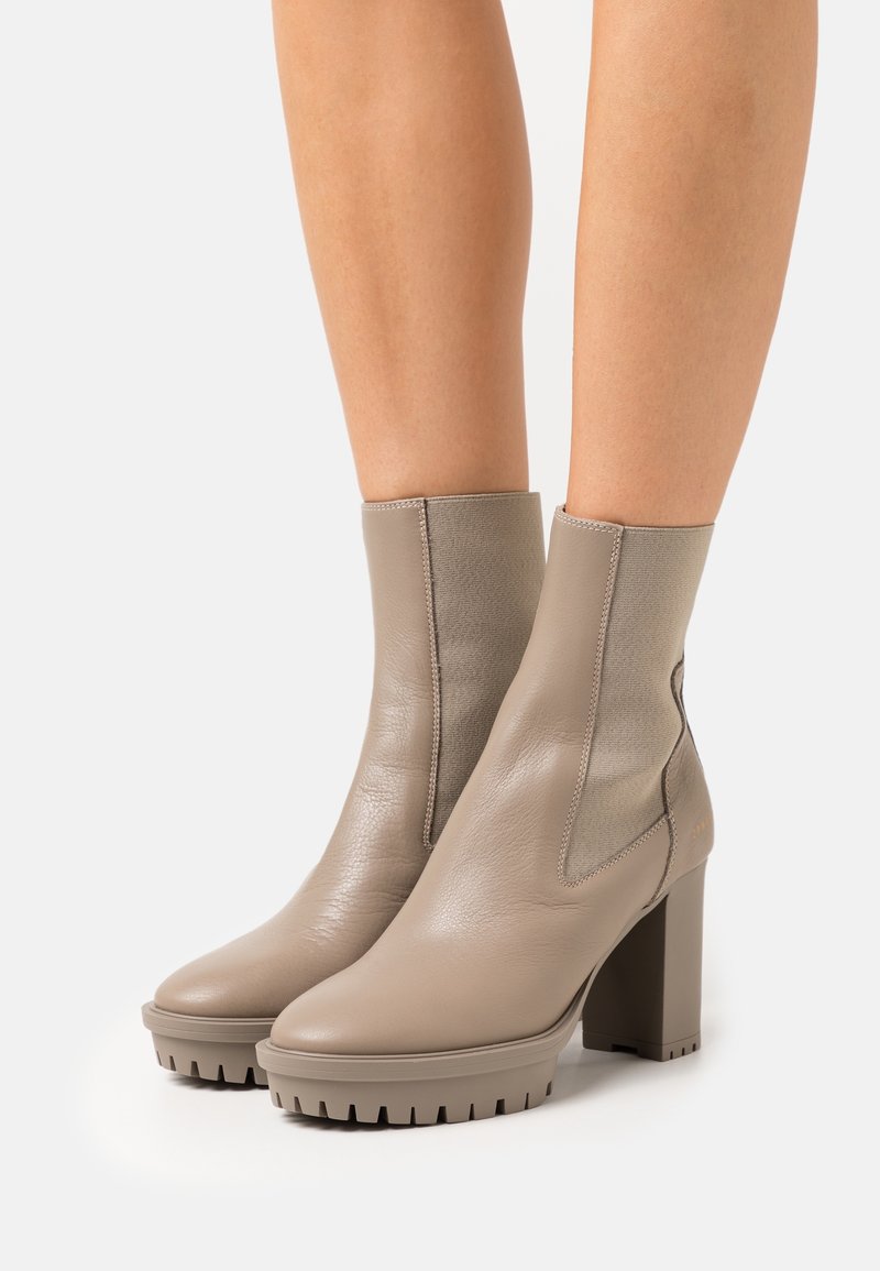 Bottes cheville en cuir taupe avec panneaux latéraux élastiques, talons carrés épais et semelles à crampons, portées sur jambes nues sur fond blanc.