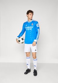 Young male soccer player in blue Real Madrid jersey, white shorts, and black cleats holding a white and black soccer ball, facing left.