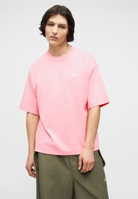 Νεαρός με μεσαίου μήκους σκούρα μαλλιά, ντυμένος με φαρδύ ροζ μπλουζάκι Adidas και χακί παντελόνι, μπροστά σε απλή φόντο.