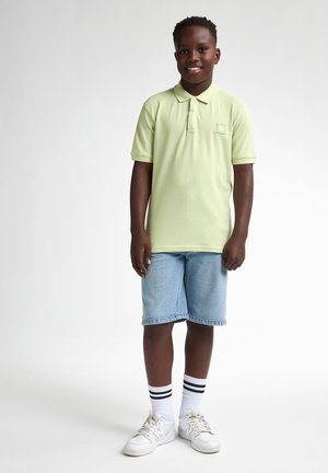 Lichtgroene poloshirt met een klein logo op de pocket, gecombineerd met blauwe denim shorts. Witte sneakers met kousen met zwarte strepen. Eenvoudige, casual stijl.