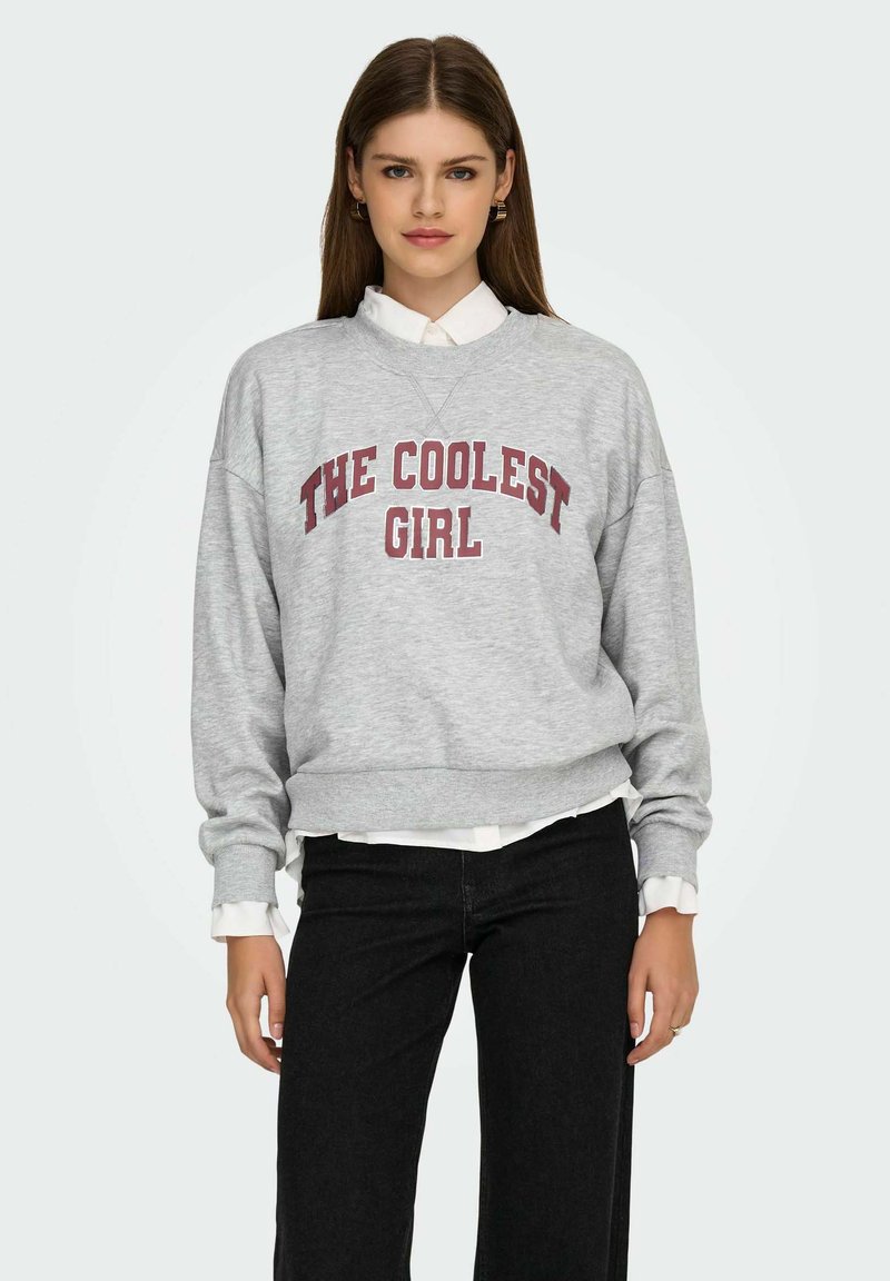 Hall halli dressipluus soonikkääristega, rinnal punase tekstiga "THE COOLEST GIRL". Kandes valge kraega särgi ja mustade pükstega.