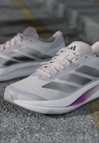 Hellrosa Sportschuhe mit grauen Adidas-Streifen, die auf strukturiertem Asphalt mit einer schwachen gelben Linie im Hintergrund liegen.