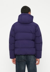 Man wearing a purple padded hooded jacket and dark jeans, standing with his back to the camera against a plain background.