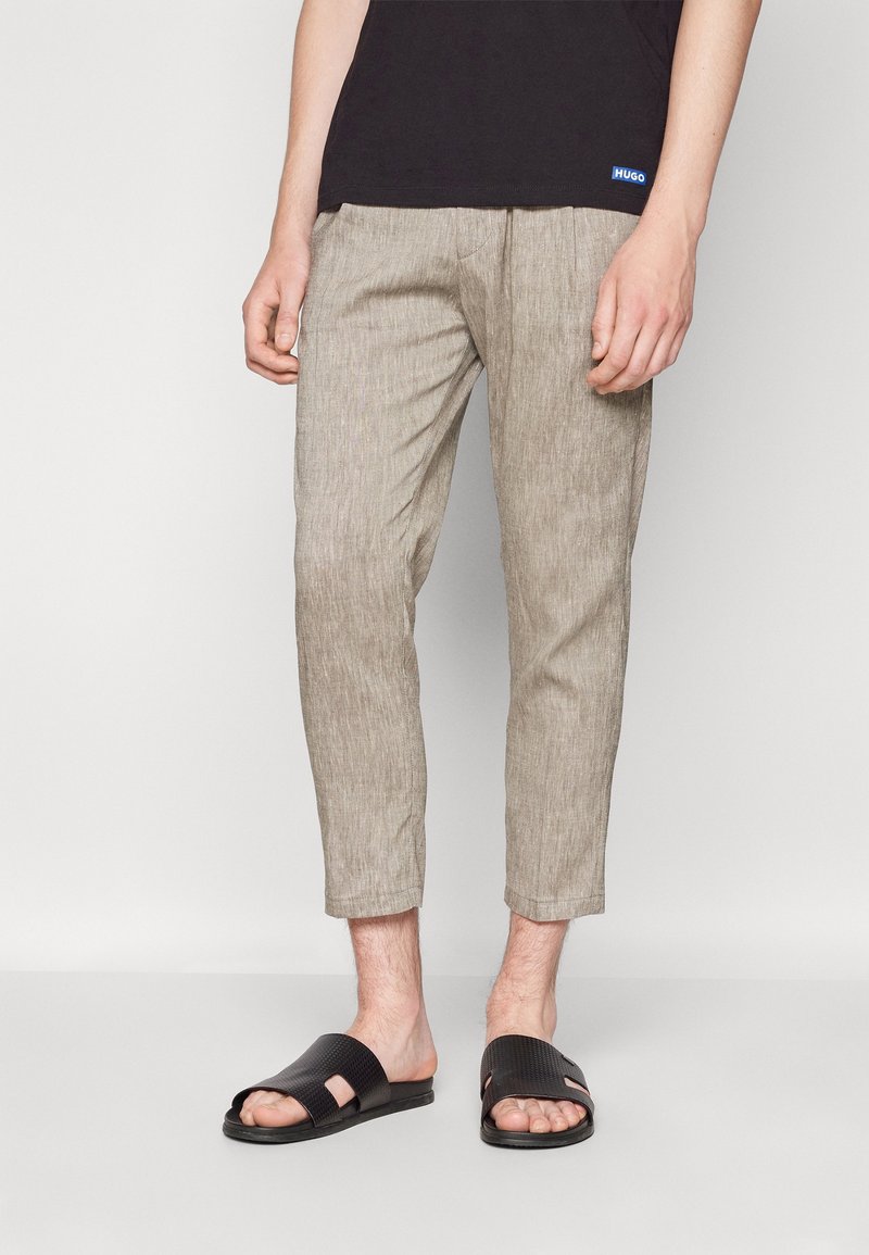 Man gedragen met gecropte beige textuurbroeken, een zwart shirt en zwarte slippers, staand tegen een effen achtergrond.