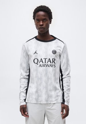 Person iført en hvid Paris Saint-Germain fodboldtrøje med lange ærmer, Qatar Airways-logo og sorte sidelister.