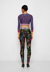 Cropped top in pizzo viola con maniche a tre quarti sopra leggings neri caratterizzati da un motivo barocco colorato e una cintura bianca con logo.