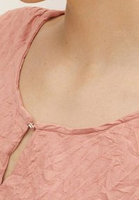 Gros plan sur une personne portant un haut rose texturé fermé par une petite attache métallique au niveau du col, dévoilant la peau et la zone de la clavicule.