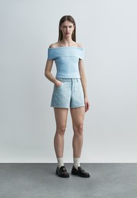 Žena stojaca s jednou rukou v kapsi, oblečená v svetlom modrom topánom bez ramienok, ozdobených džínsových šortkách, bielych ponožkách a čiernych mokasínach.