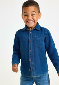 Camicia in denim blu medio, caratterizzata da maniche lunghe, una tasca sul petto e bottoni marroni. Tessuto testurizzato con dettagli di cucitura visibili.
