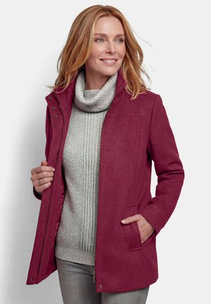 Frau trägt einen burgunderfarbenen Mantel über einem grauen, gestrickten Rollkragenpullover und grauen Hosen, lächelt und schaut zur Seite.