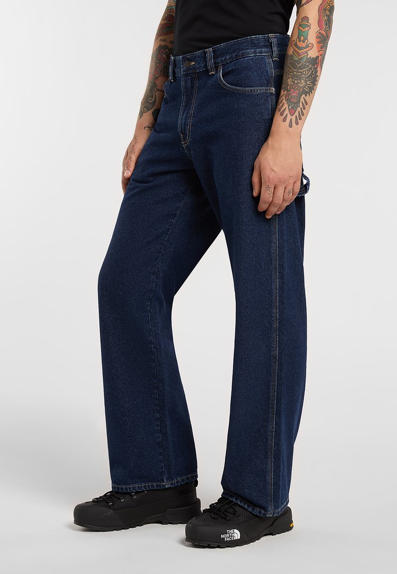 Person iført mørkeblå jeans med lige ben, sorte sneakers med The North Face-logo og en sort top, der viser tatoverede arme.