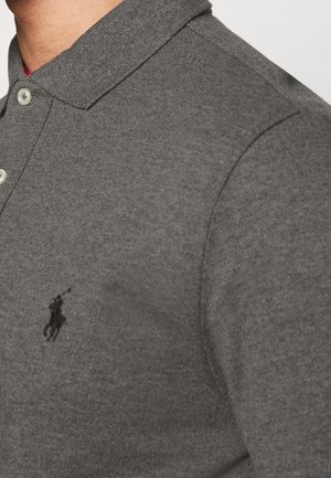 Gros plan d'une personne portant un polo gris avec un logo de joueur de polo brodé en noir sur la poitrine et deux boutons blancs près du col.