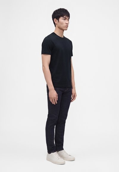 Νέος άνδρας στέκεται, φορόντας απλό μαύρο T-shirt, σκούρα τζιν και λευκά αθλητικά παπούτσια σε ουδέτερο φόντο.